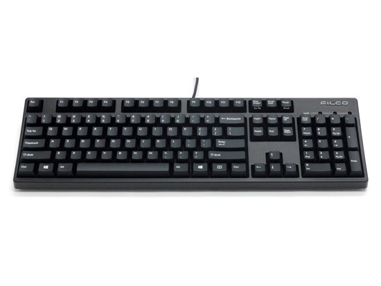 Gambar keyboard Filco Majestouch 2 di atas meja dengan monitor yang menampilkan baris kode, menekankan desainnya yang bersih dan utilitarian serta lingkungan kerja seorang programmer yang terfokus.