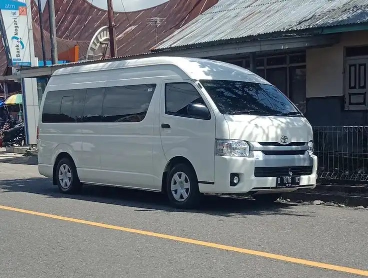 Gambar Toyota Hiace Commuter diparkir di depan hotel atau bangunan komersial dekat Living World Denpasar, menonjolkan keluasannya dan kegunaannya untuk transportasi penumpang.