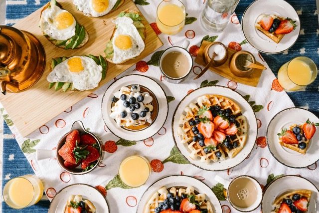 Meja penuh hidangan brunch klasik seperti eggs benedict, waffle, dan kopi.