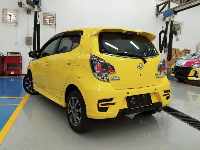 Gambar Daihatsu Ayla kuning terparkir rapi di jalan sempit di kota yang ramai, menonjolkan ukuran kompak dan kesesuaian perkotaannya, dengan latar belakang samar Jakarta Timur.