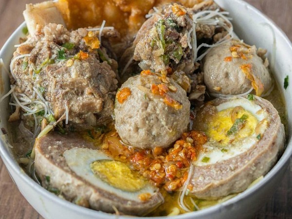 Semangkuk bakso panas dengan kuah yang mengepulkan asap, di latar belakang terlihat rintik hujan.