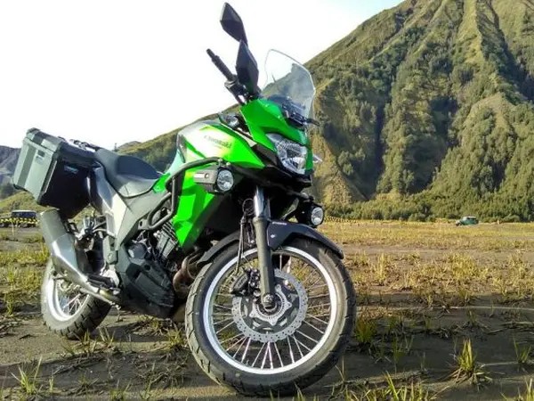 Gambar Kawasaki Versys-X 250 diparkir di luar jalan dengan lanskap Indonesia yang indah (misalnya, pegunungan atau jalan tanah yang mengarah ke hutan) di latar belakang, menekankan kemampuan petualang