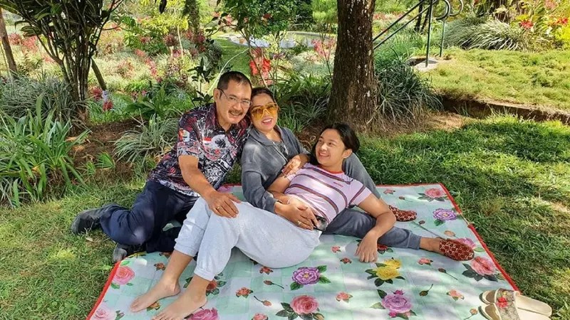 Seseorang sedang duduk santai di bangku taman yang dikelilingi pepohonan rindang dan danau kecil.