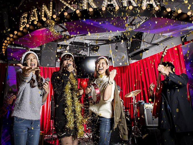 Seseorang bernyanyi di panggung kecil diiringi live band di The Stage Karaoke & Bar.