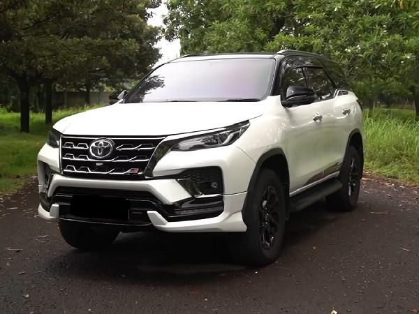 Gambar Toyota Fortuner putih terparkir di jalan pemandangan dengan keluarga menikmati pemandangan, menonjolkan sifat tangguh namun ramah keluarga, dengan latar belakang samar Surabaya.