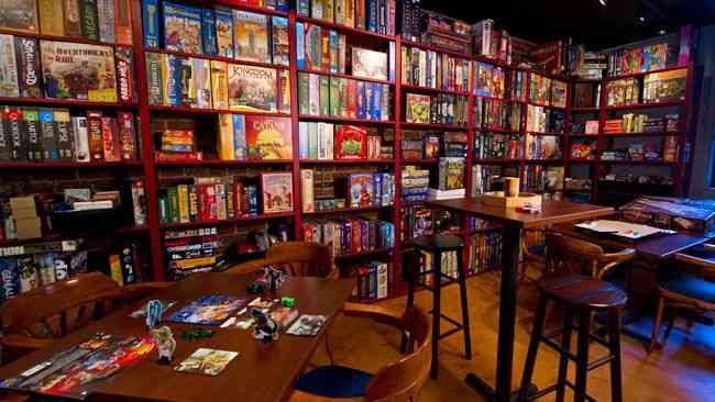 Orang-orang sedang bermain board game di meja cafe dengan banyak game box di rak belakang.