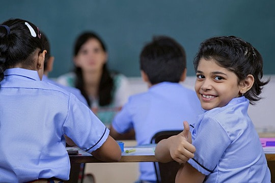 Gambar seorang guru tersenyum sedang mengajar di depan kelas yang berisi siswa-siswi yang antusias.