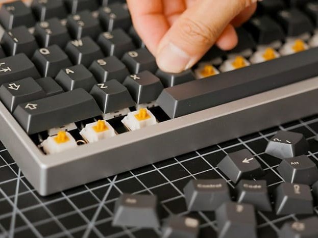 Gambar komponen keyboard mekanik yang dibongkar (PCB, pelat, sakelar, keycap) ditata rapi, dengan tangan sedang merakit salah satu bagian, menggambarkan proses kustomisasi.