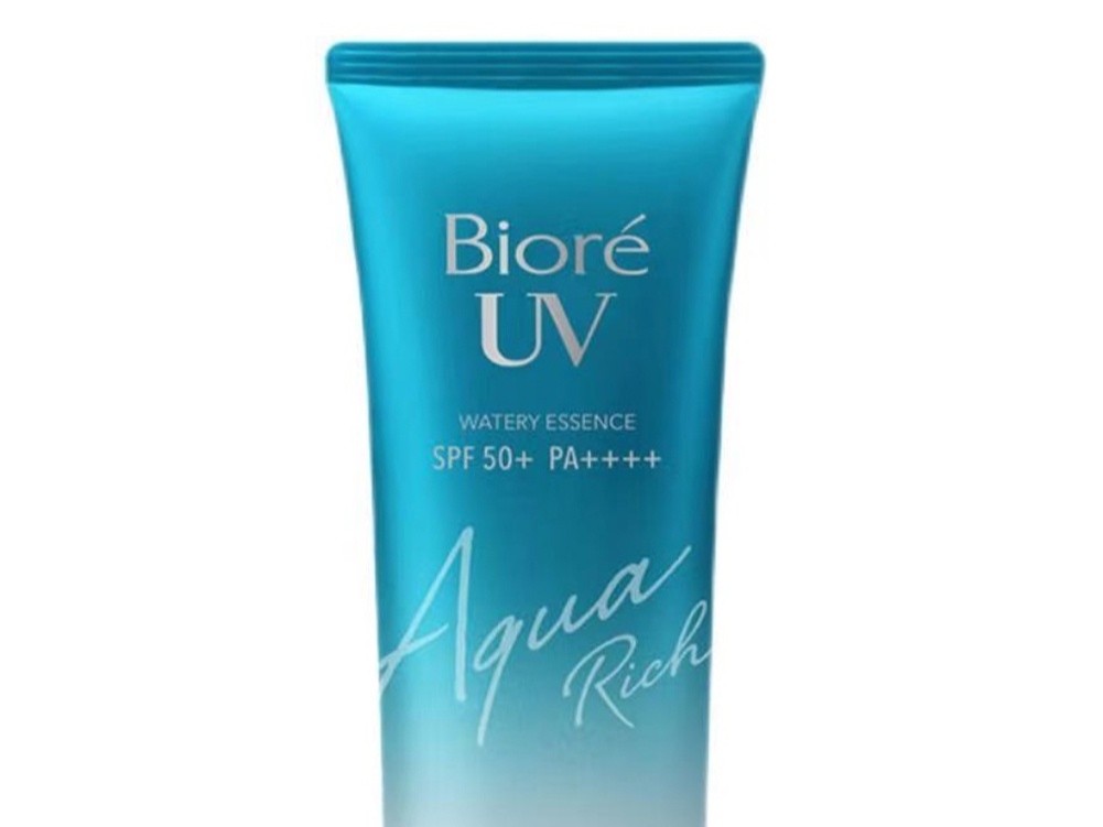 Gambar botol Biore UV Aqua Rich Watery Essence di samping tekstur cairnya yang menyebar di kulit, menonjolkan ringannya dan cepat meresap, dengan latar belakang samar Jakarta Selatan.