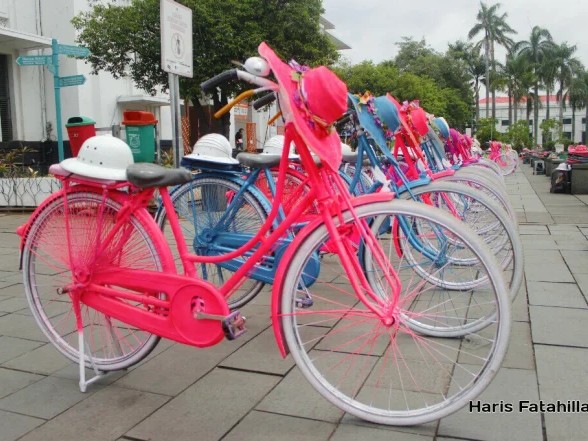 Gambar sepeda fixie United merah yang stylish terparkir di lingkungan perkotaan yang estetis, mungkin di dinding grafiti atau bangunan tua, menonjolkan gaya uniknya, dengan latar belakang samar Kota T