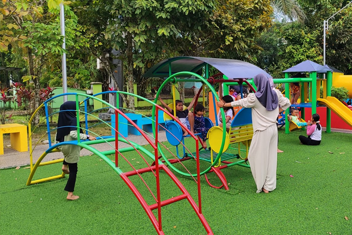 Anak-anak dan remaja bermain di wahana permainan outdoor dengan pemandangan hijau dan langit cerah.