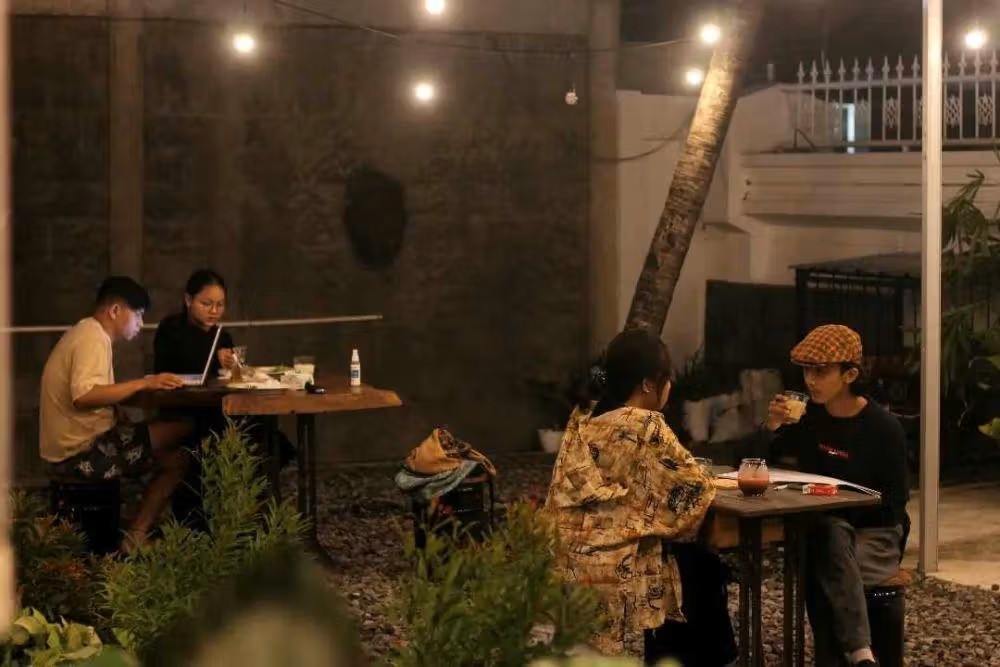 Interior cafe dengan pencahayaan temaram, sofa nyaman, dan beberapa orang sedang nongkrong di malam hari.