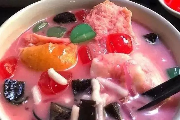 Semangkuk es campur atau es teler penuh dengan warna-warni isian, ada es serut dan siraman sirup, suasana menyegarkan.