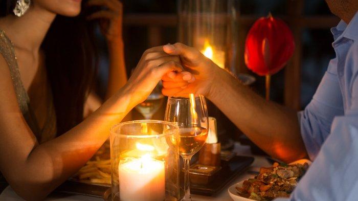 Pasangan sedang makan malam romantis di restoran dengan pencahayaan temaram dan lilin.