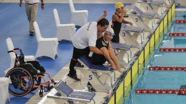 Seseorang dengan kursi roda di tepi kolam renang yang dilengkapi ramp akses khusus.