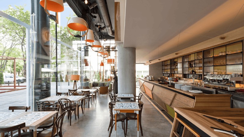 Interior coffee shop yang estetik dengan pencahayaan alami yang masuk melalui jendela besar, terlihat beberapa spot duduk yang nyaman.