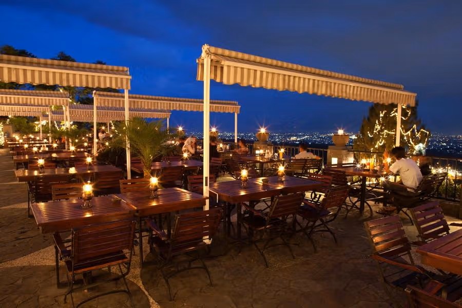 Pasangan menikmati makan malam romantis di rooftop restaurant dengan pemandangan kota Bandung di malam hari.