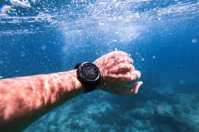 Jam tangan diver profesional di pergelangan tangan, dengan latar belakang air laut yang jernih dan karang.