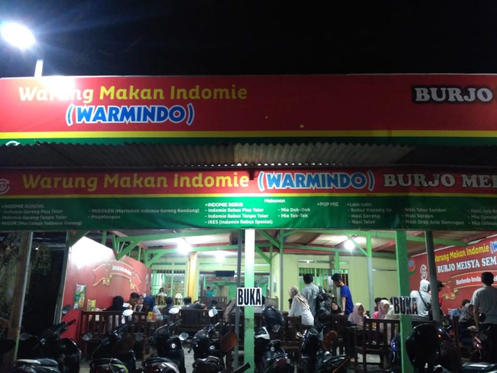 Suasana Warmindo ramai di malam hari dengan mahasiswa yang makan mie instan dan nongkrong.