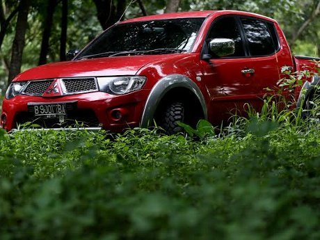 Gambar Mitsubishi Triton merah melaju di jalan tanah yang kasar dengan latar belakang lokasi konstruksi atau hutan, menonjolkan daya tahan dan kegunaannya, dengan latar belakang samar Samarinda Kalima