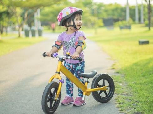 Gambar sepeda balance bike Balance Bike Indonesia (BiBi) berwarna-warni dengan balita bahagia meluncur di taman, dengan orang tua bersorak, menonjolkan kesenangan dan pembelajaran, dengan latar belaka