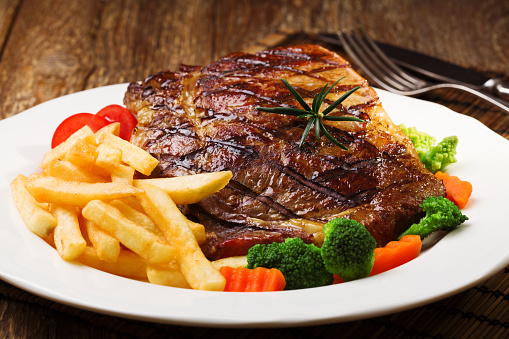 Piring berisi steak daging sapi di hot plate dengan kentang goreng dan sayuran hijau.