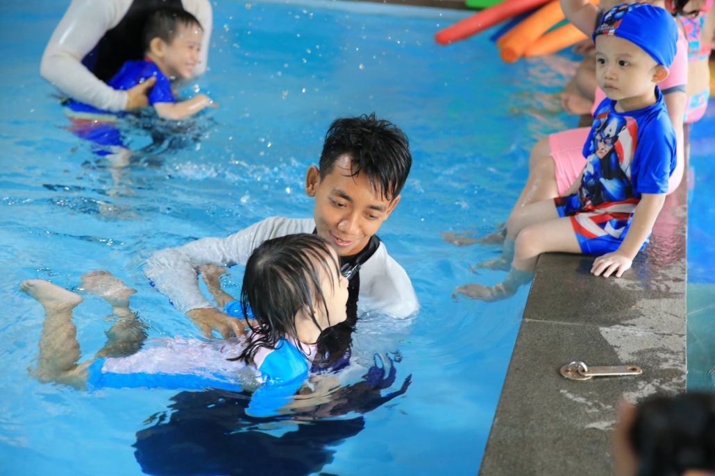 Instruktur renang sedang mengajari anak kecil berenang di kolam dangkal.