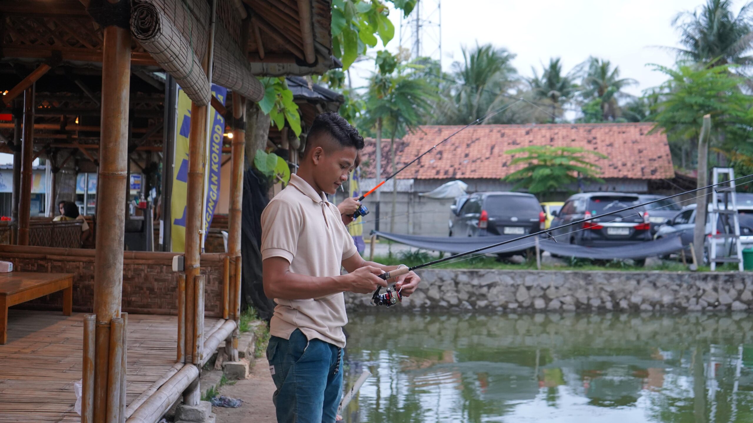 Orang sedang memancing di kolam pemancingan dengan saung-saung di sekelilingnya, suasana tenang.