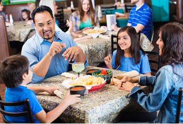 Keluarga sedang makan di restoran modern dengan area bermain anak yang terlihat di latar belakang.