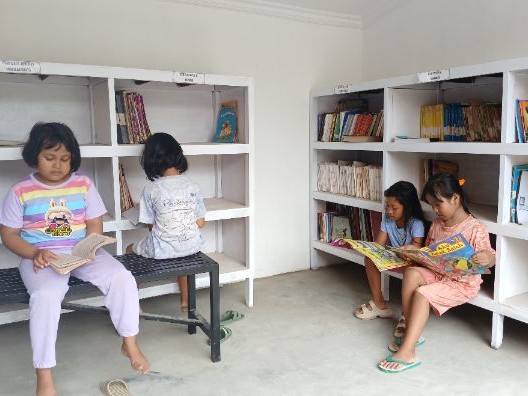 Gambar interior perpustakaan desa kecil dengan warga lokal membaca.