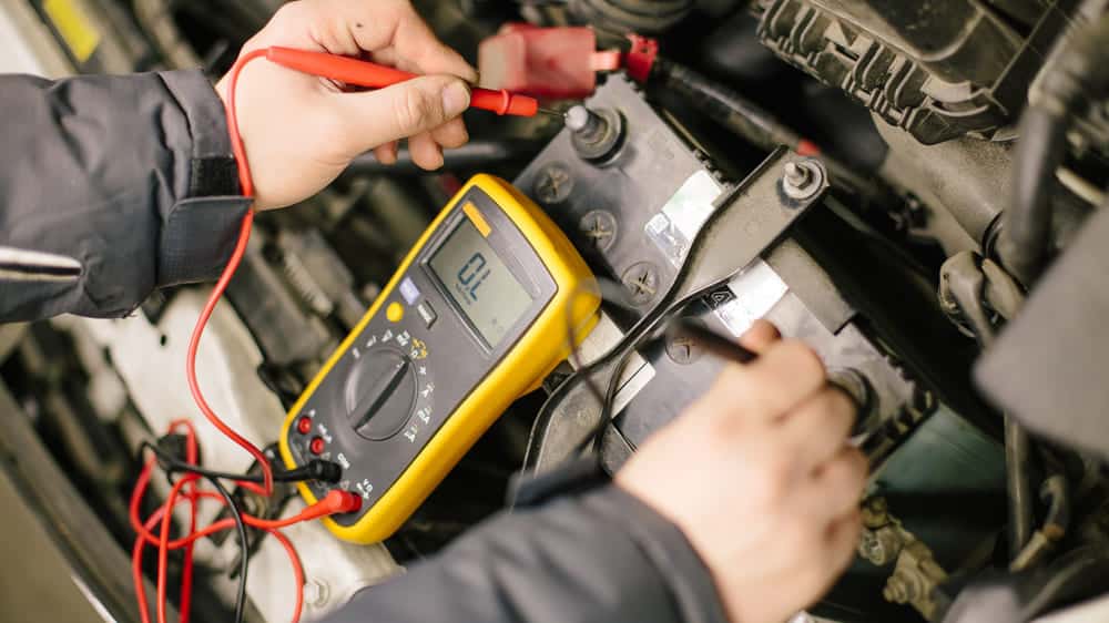 Mekanik sedang memeriksa terminal aki mobil dengan alat pengukur tegangan.