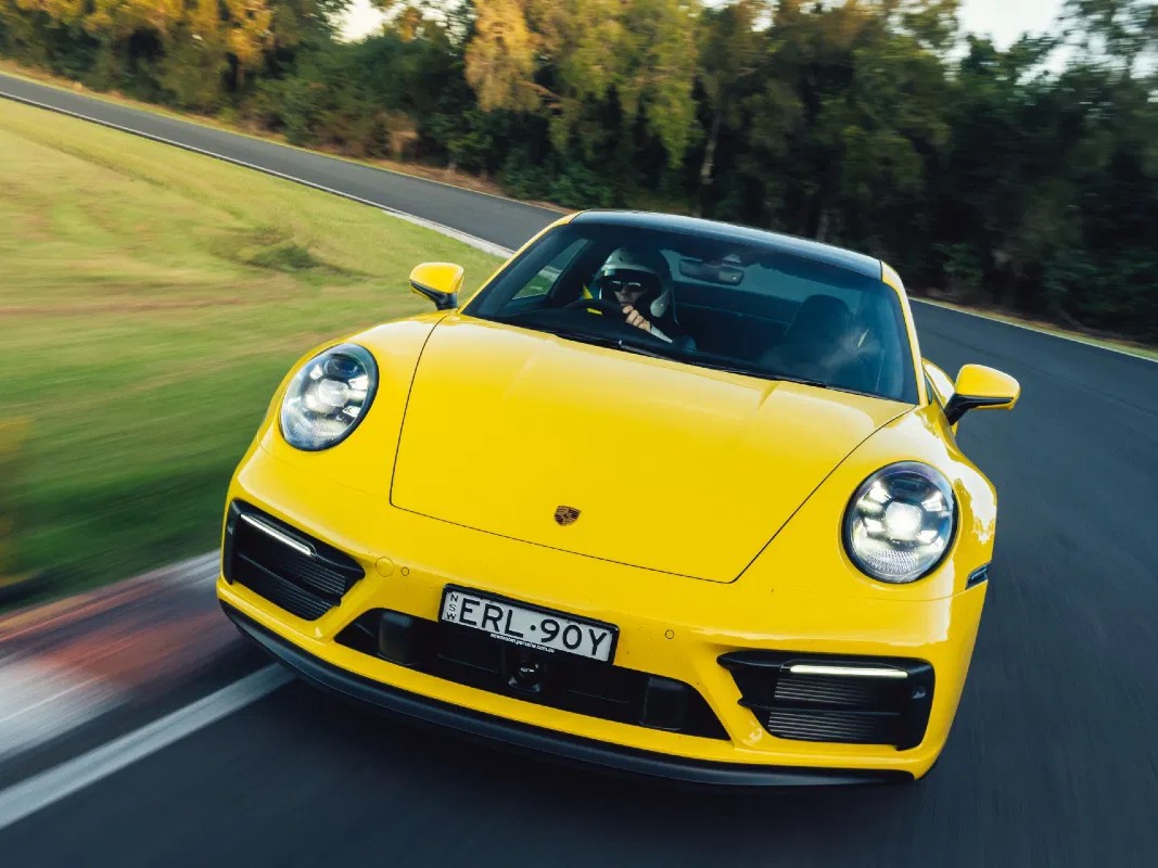 Gambar Porsche 911 Carrera S kuning melaju di lintasan balap atau jalan gunung berkelok-kelok, menonjolkan performa dan desain rampingnya, dengan latar belakang samar Jakarta Selatan.