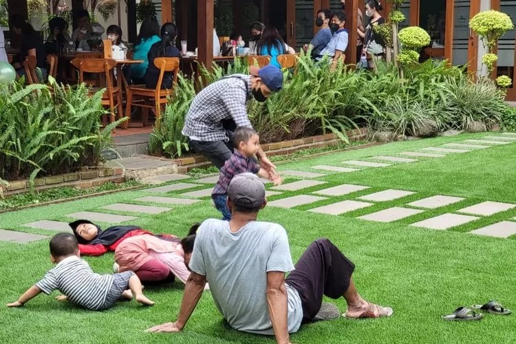 Keluarga sedang makan di area outdoor restoran yang luas, dengan anak-anak bermain di area playground.