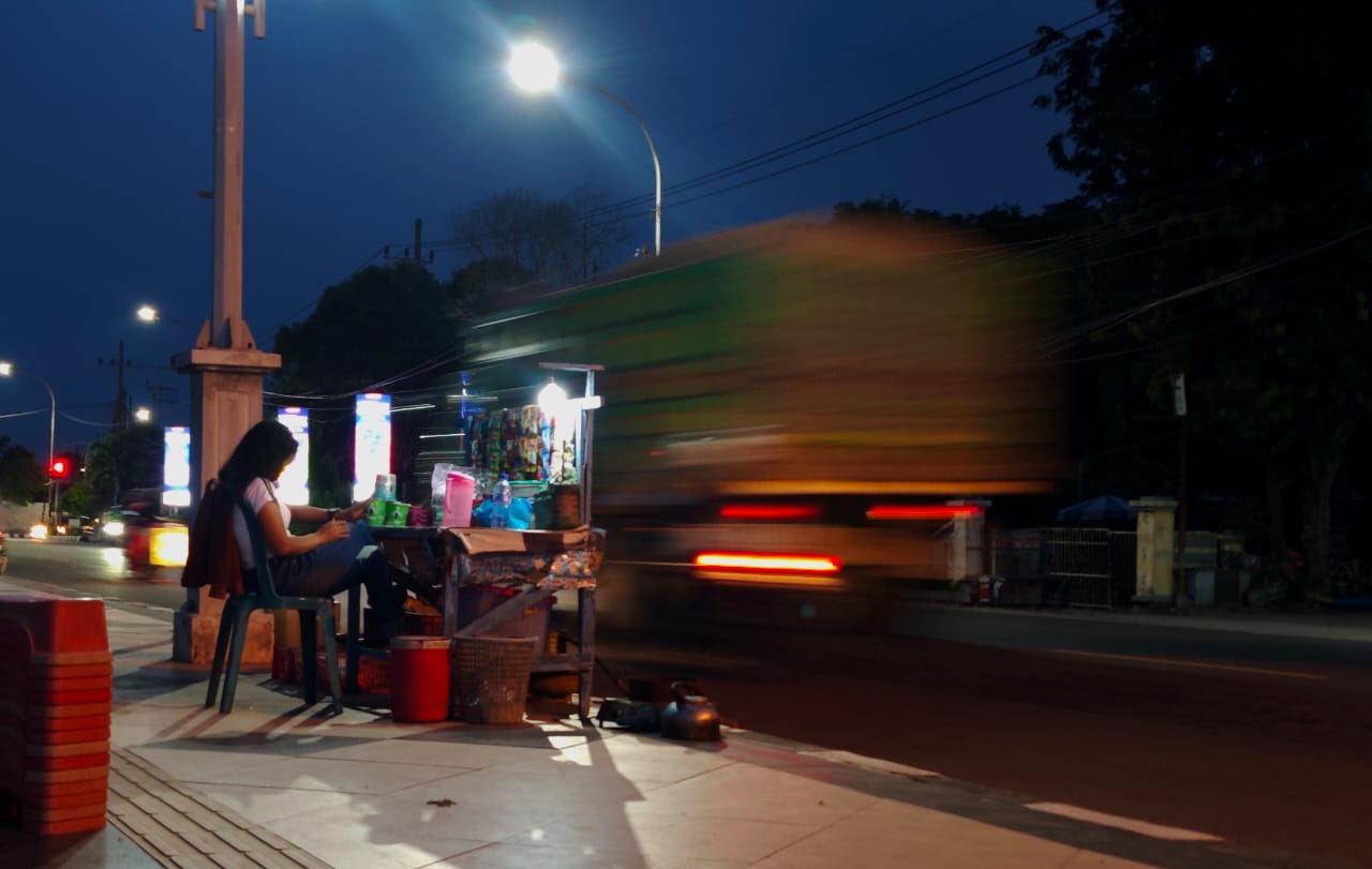 Suasana warung makan di pinggir jalan yang ramai di malam hari dengan cahaya lampu.