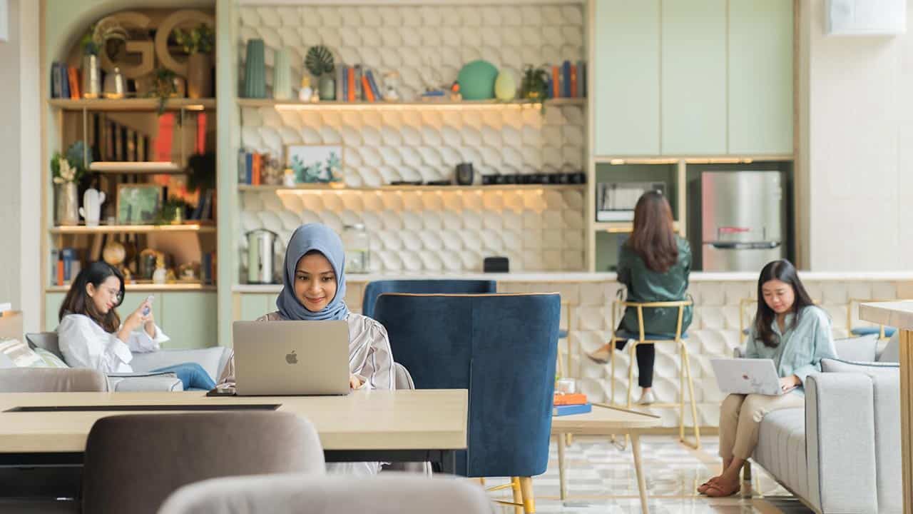 Orang-orang sedang bekerja dengan laptop di meja panjang sebuah co-working space yang modern.