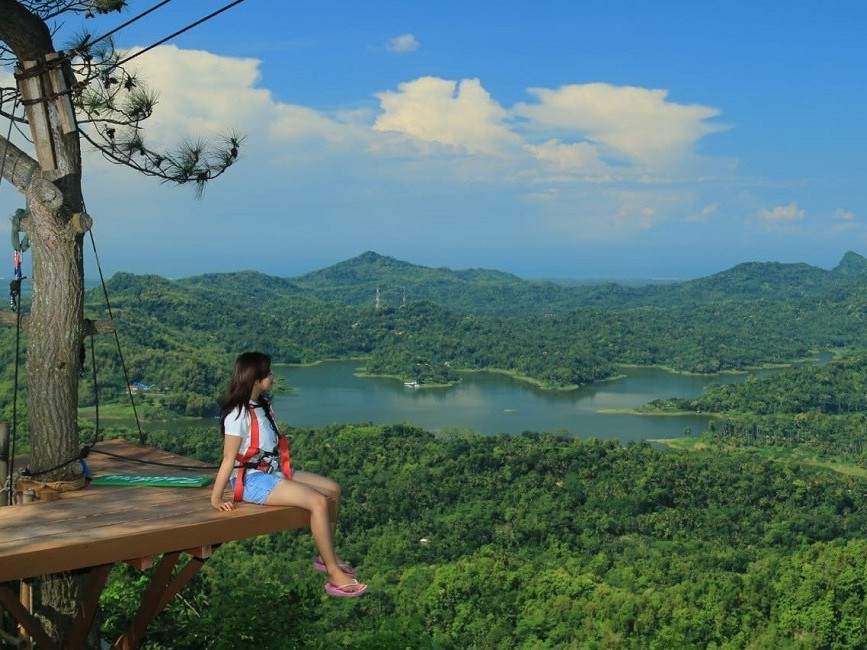 Gambar seseorang berdiri di platform kayu melihat danau dan bukit di Kalibiru.