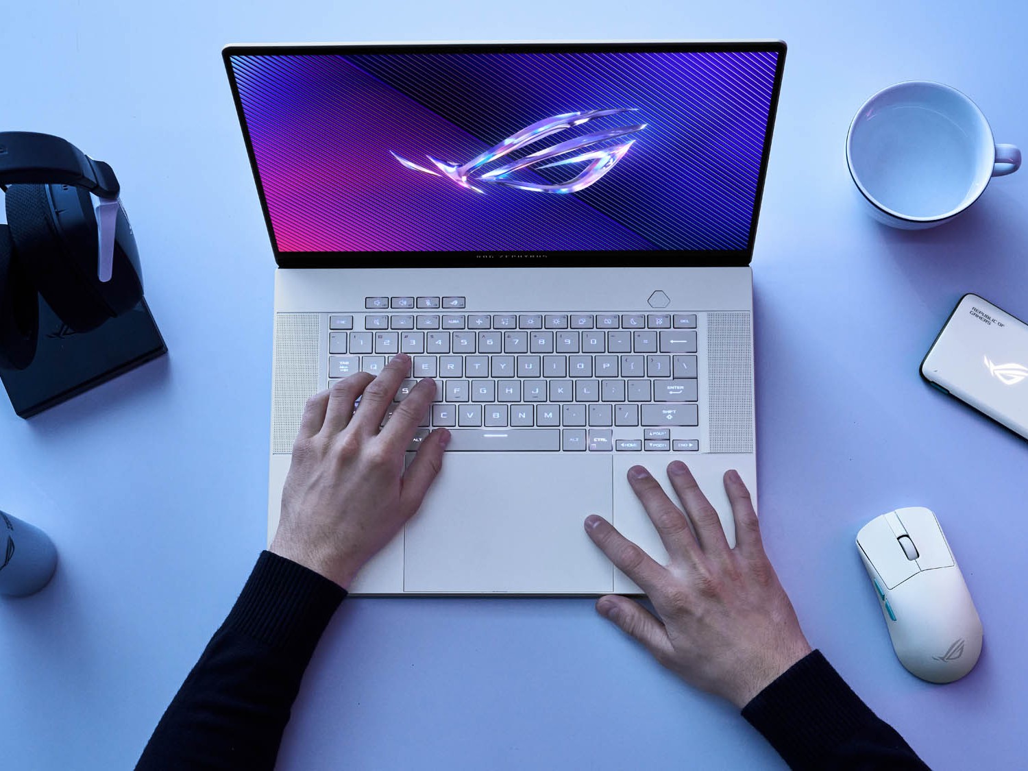 Gambar ASUS ROG Zephyrus G14/G16 di meja gaming dengan pencahayaan RGB, dikelilingi periferal gaming, terletak di zona gaming atau dekat Mangga Dua Mall, Jakarta Utara.