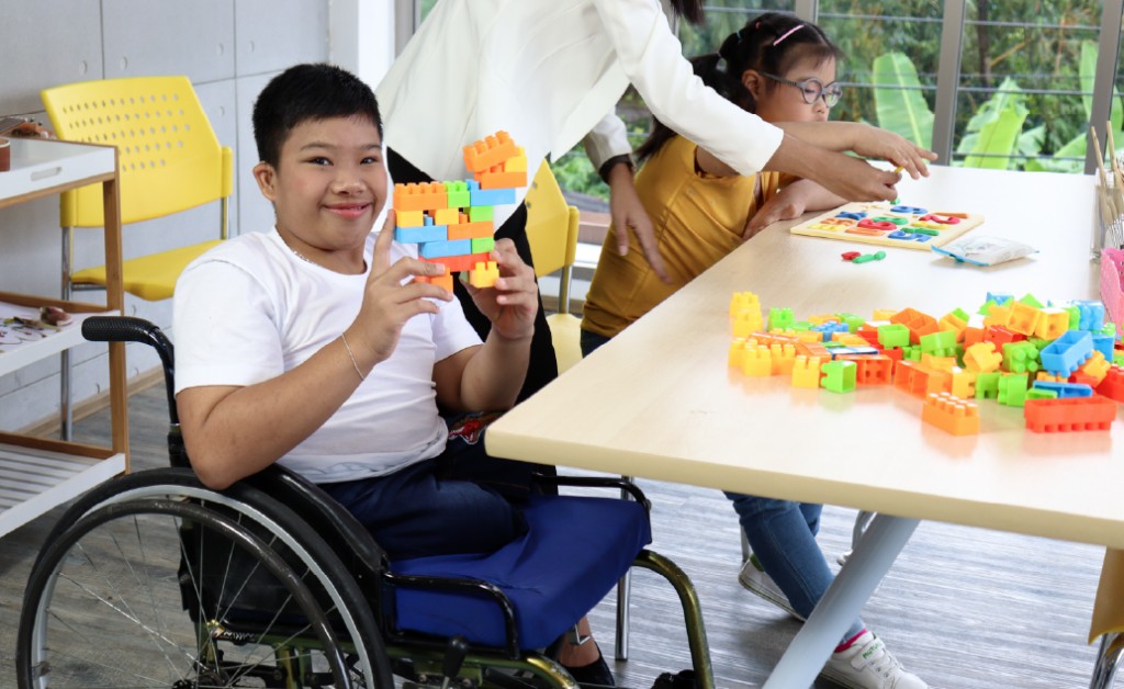 Gambar seorang guru berinteraksi dengan siswa menggunakan kursi roda di kelas, menunjukkan dukungan dan inklusivitas.