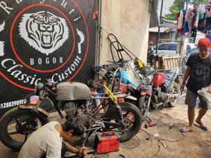 Gambar bengkel kustomisasi motor dengan berbagai motor custom dalam tahap penyelesaian yang berbeda, serta peralatan/suku cadang yang tertata rapi.