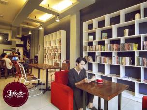 Orang sedang membaca buku di kafe dengan rak buku besar dan suasana tenang.