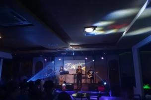 Panggung live music akustik di sebuah kafe dengan penonton yang menikmati alunan musik.