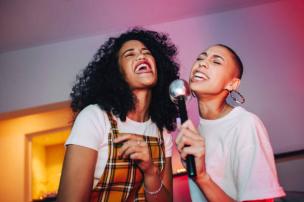 Pasangan muda sedang bernyanyi dengan ceria di ruangan karaoke modern The Voice Karaoke dengan pencahayaan warna-warni.