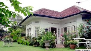 Gambar rumah bergaya kolonial dengan teras luas dan taman di daerah pegunungan yang sejuk.