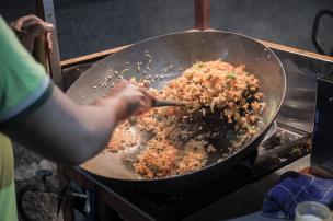 Nasi goreng sedang dimasak di wajan besar dengan api membara, mengeluarkan aroma asap.