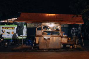 Warung nasi goreng kaki lima di malam hari, dengan asap mengepul dari wajan, dan beberapa pembeli mengantre.