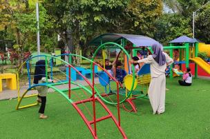 Anak-anak dan remaja bermain di wahana permainan outdoor dengan pemandangan hijau dan langit cerah.