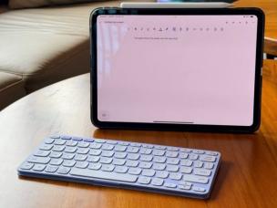 Gambar keyboard Logitech Keys-to-Go yang tipis dan ringan di samping tablet di lingkungan kafe, menekankan portabilitas dan kemudahan penggunaannya untuk bekerja jarak jauh.