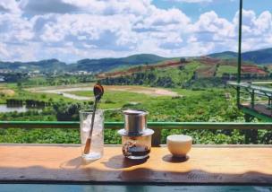 Orang-orang bersantai di kafe outdoor dengan pemandangan perbukitan hijau dan langit biru.