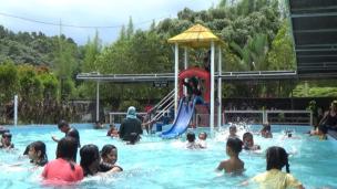 Kolam renang umum yang ramai dengan orang-orang berenang dan bersantai di siang hari.