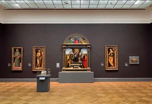 Interior galeri seni dengan beberapa lukisan modern dipajang di dinding putih dan pengunjung sedang mengamati.